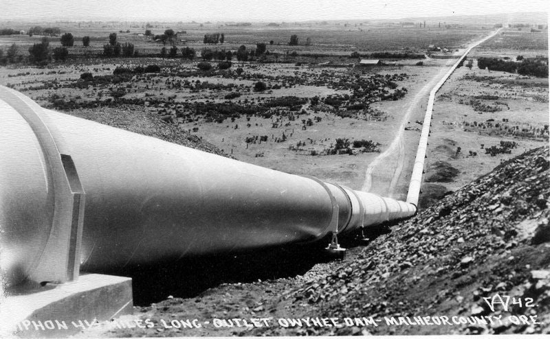 An old black-and-white photo of a large siphon pipe from the Owyhee Dam in Malheur County, Oregon, overlooking a landscape.