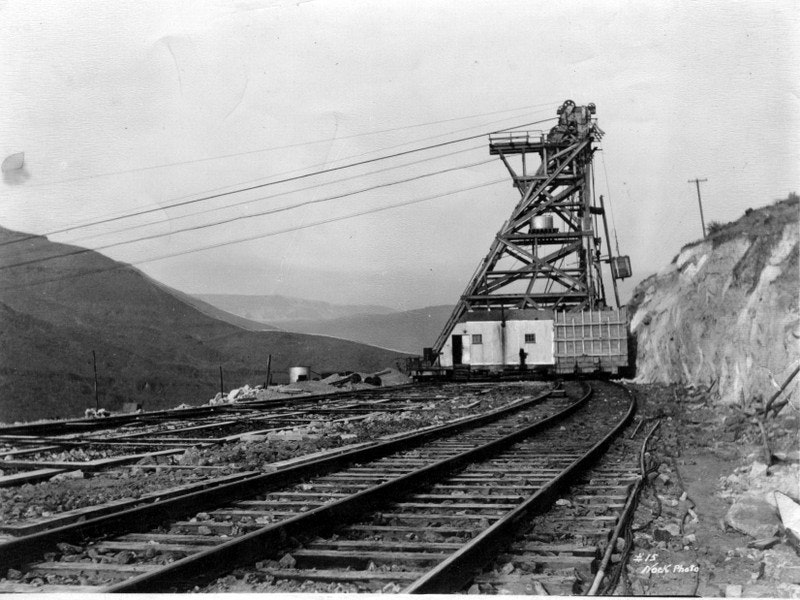 The image shows a historic mining operation with a large hoisting tower and railroad tracks in a mountainous landscape.