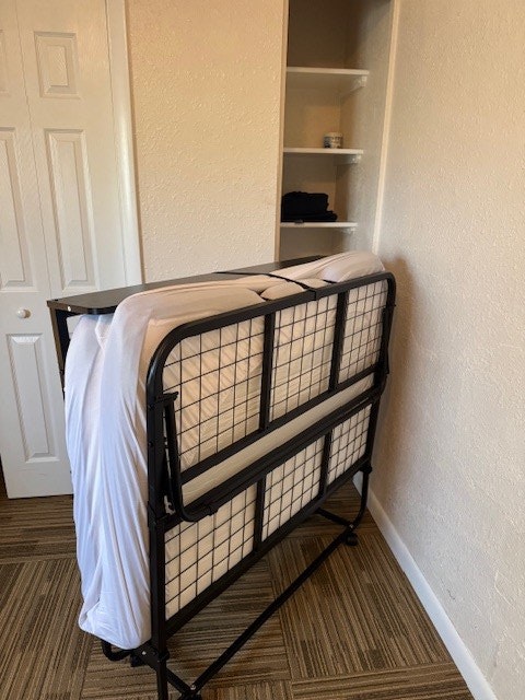 A foldable bed is propped up against a wall in a room with a small closet and carpeted floor.