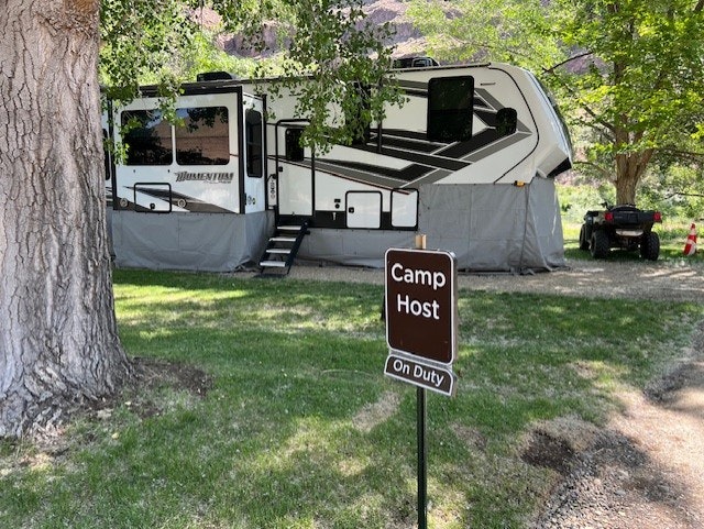 A camper labeled "Camp Host On Duty" is parked among trees with a nearby ATV.