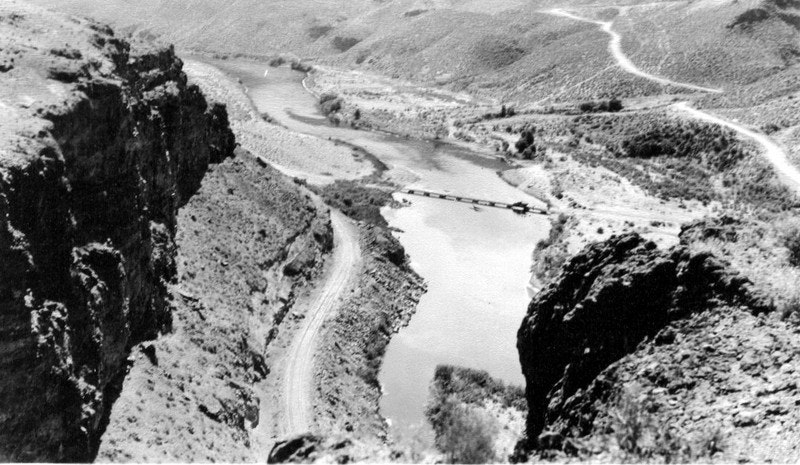 A black and white landscape featuring a winding river, rocky cliffs, and a bridge in a remote, rugged terrain.