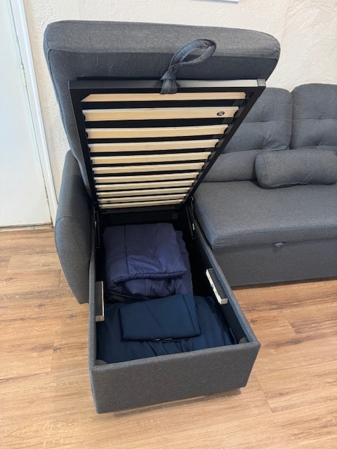 A grey storage sofa with an open lid revealing blankets inside, on a wooden floor.