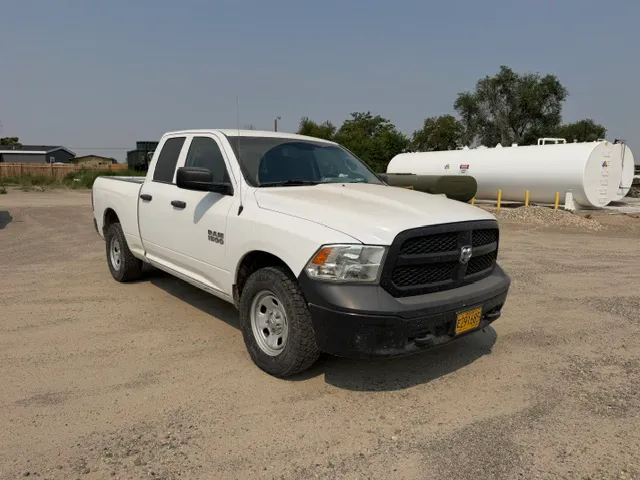 A white pickup truck parked on a gravel lot, with a tank and trees in the background.