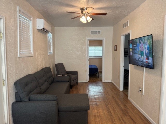 Living room with gray sofas, wall-mounted TV, ceiling fan, wooden floor, and a view into a bedroom.