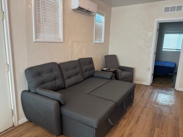 A room with a gray sofa bed, armchair, hardwood floor, wall-mounted air conditioner, and doorway leading to another room with a bed.