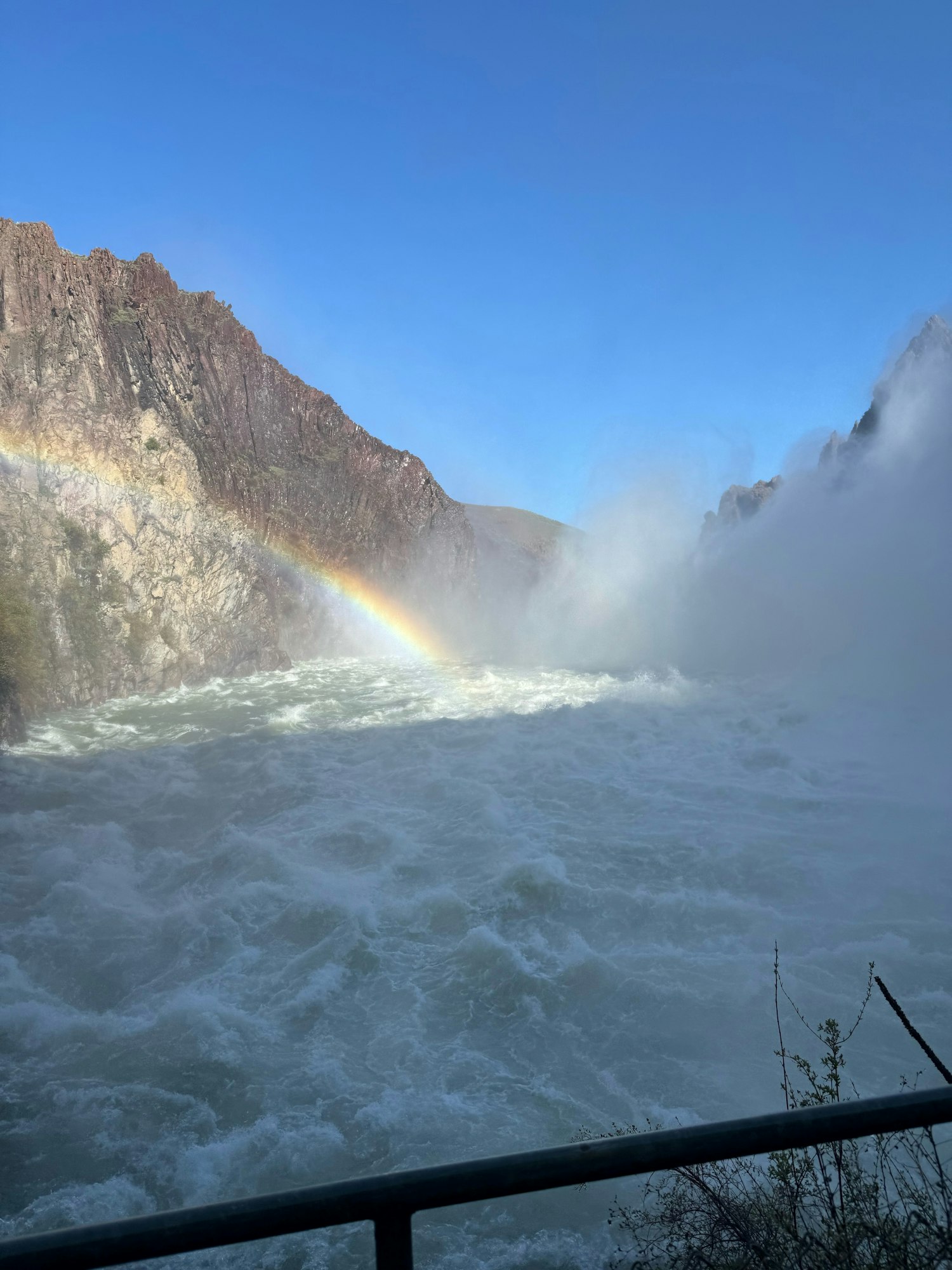 Rushing river with mist and a partial rainbow against mountainous terrain.