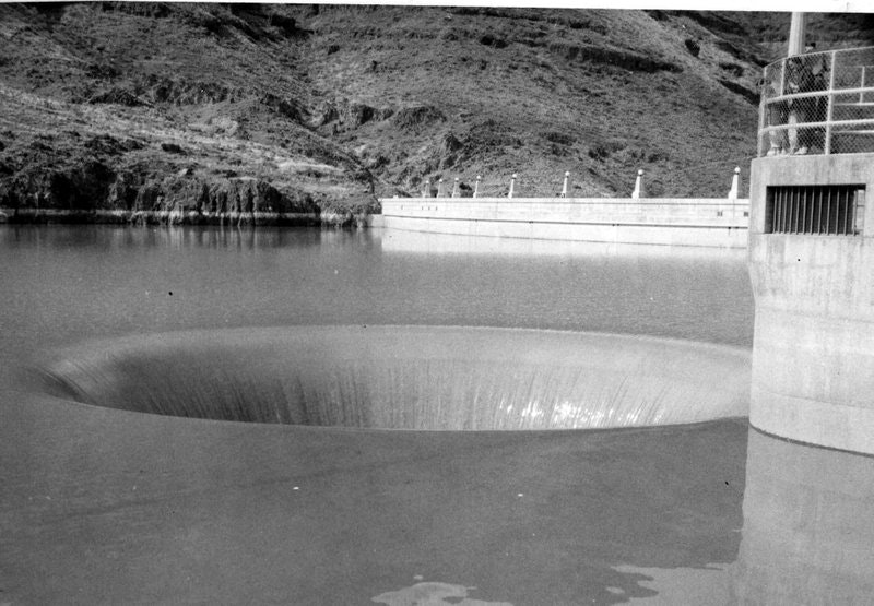 The image shows a large circular spillway or drain structure in a reservoir, with surrounding hills in the background.
