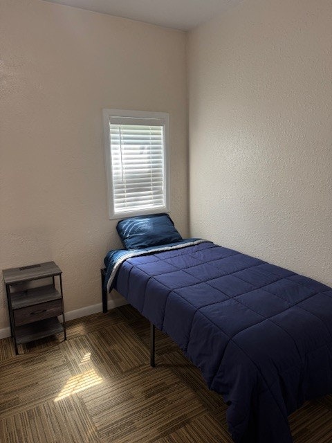 A simple bedroom with a single bed, blue bedding, small nightstand, and a window with blinds.