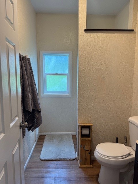 A small bathroom with a toilet, towels on a rack, a window, a bath mat, and a small cabinet holding a toilet paper roll.