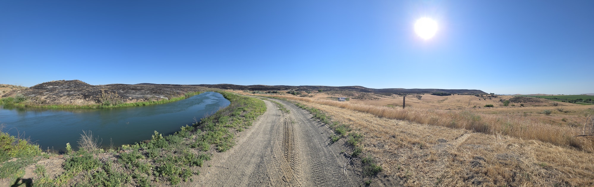 A panoramic view of a dry landscape with a river, hills, and clear blue sky, featuring a sun shining brightly above.