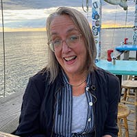 May contain: face, head, person, photography, portrait, adult, female, woman, accessories, glasses, clothing, coat, jacket, dining table, furniture, table, jewelry, necklace, water, and waterfront