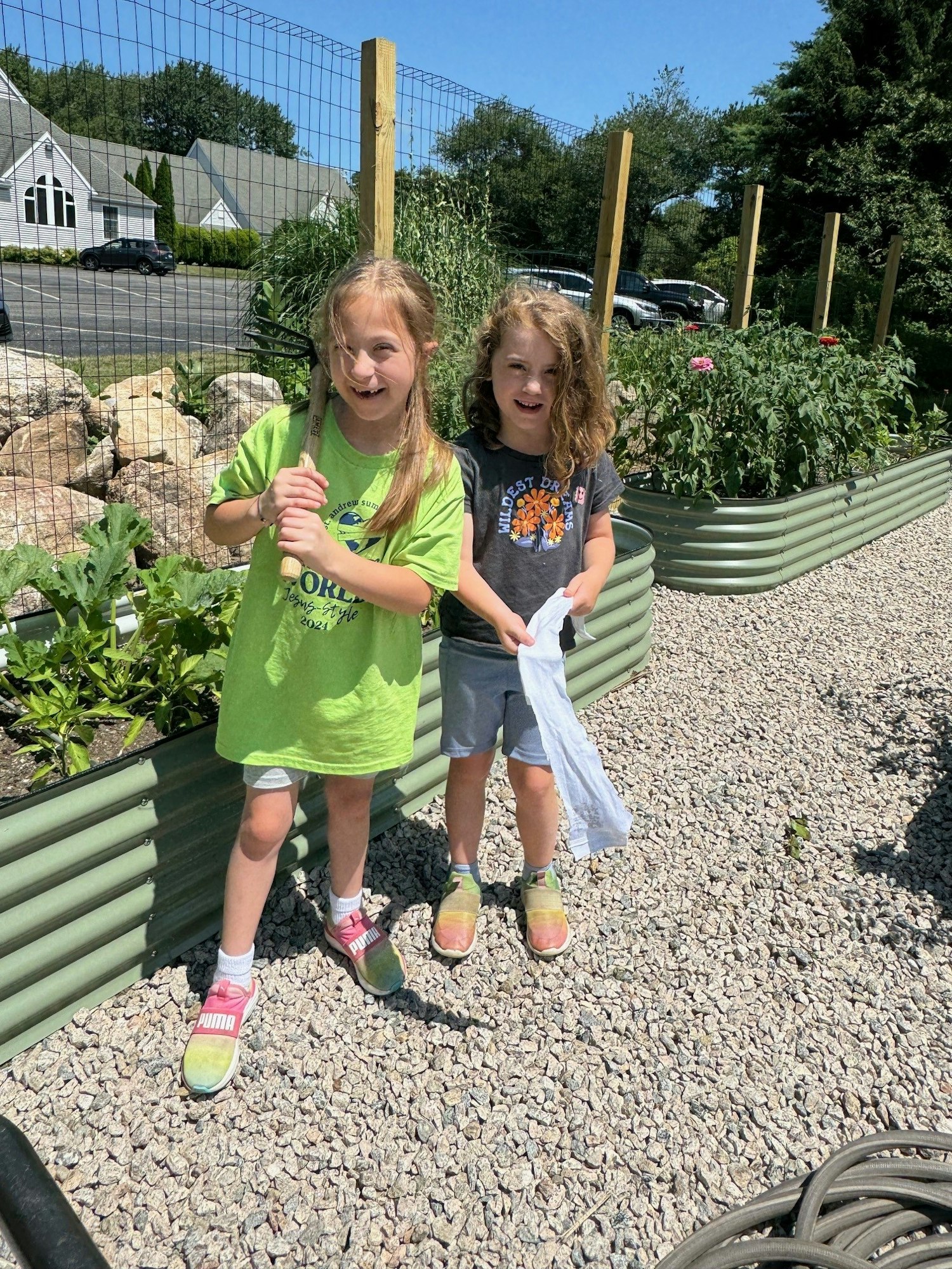 Two children standing in a garden, one holding a gardening fork, with raised beds and plants in the background.