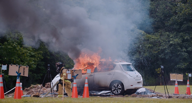 A burning car with smoke, surrounded by traffic cones and firefighting personnel outside.