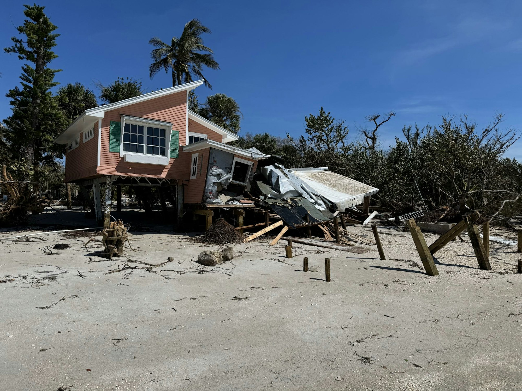 Damaged house with debris, possibly from a natural disaster, surrounded by trees under blue sky.