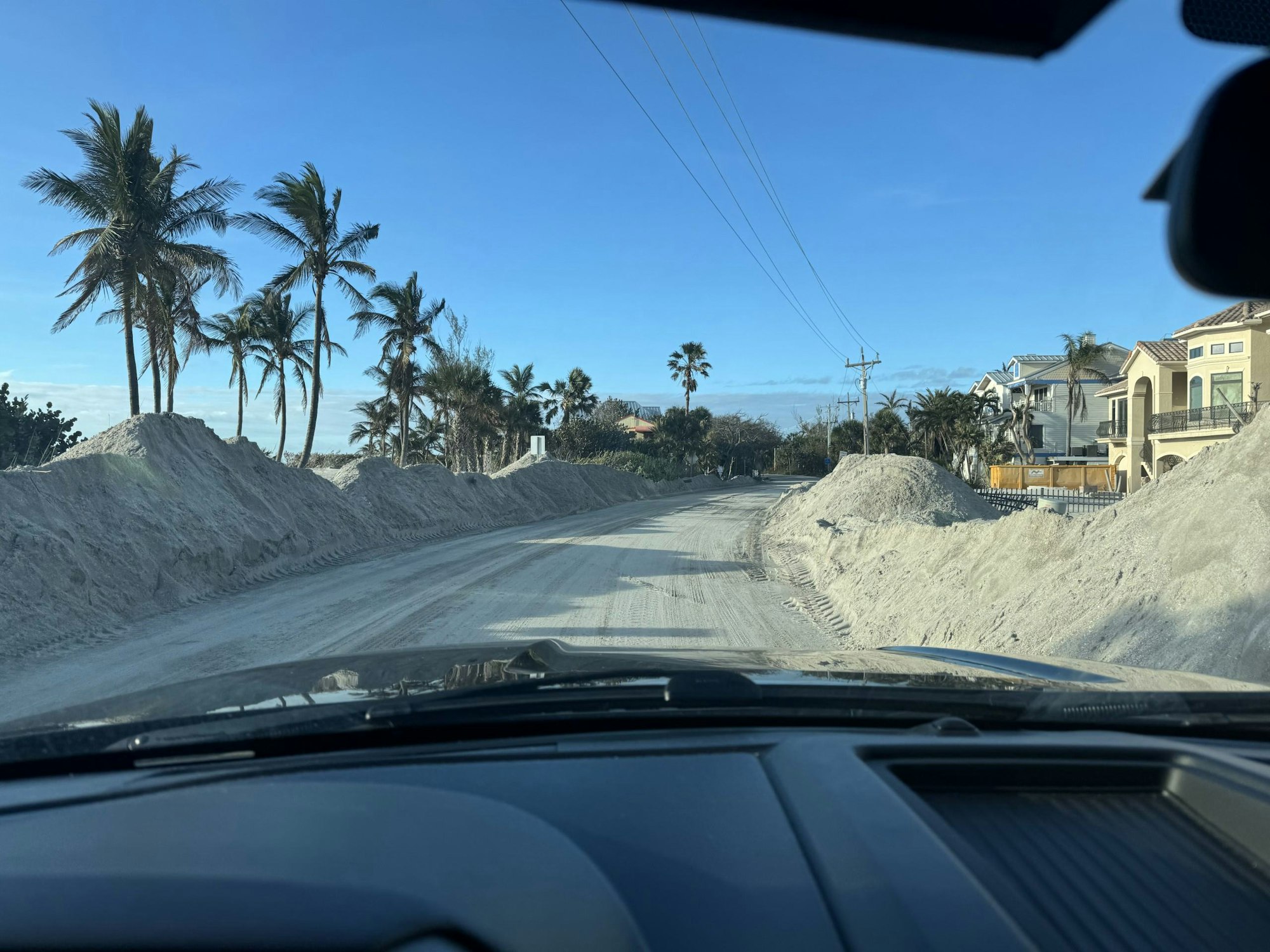 View from a car showing a sandy road lined with palm trees and houses, large sand piles on the side.