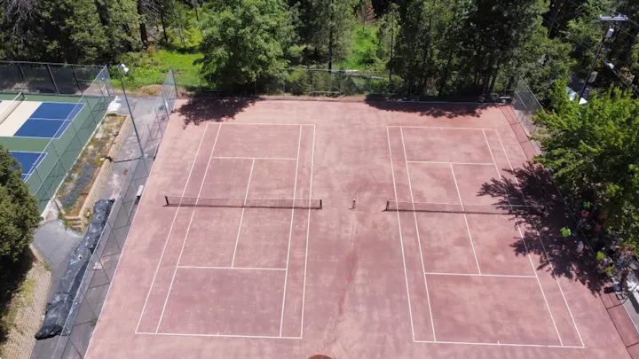 Two tennis courts surrounded by trees, adjacent to a pickleball court.