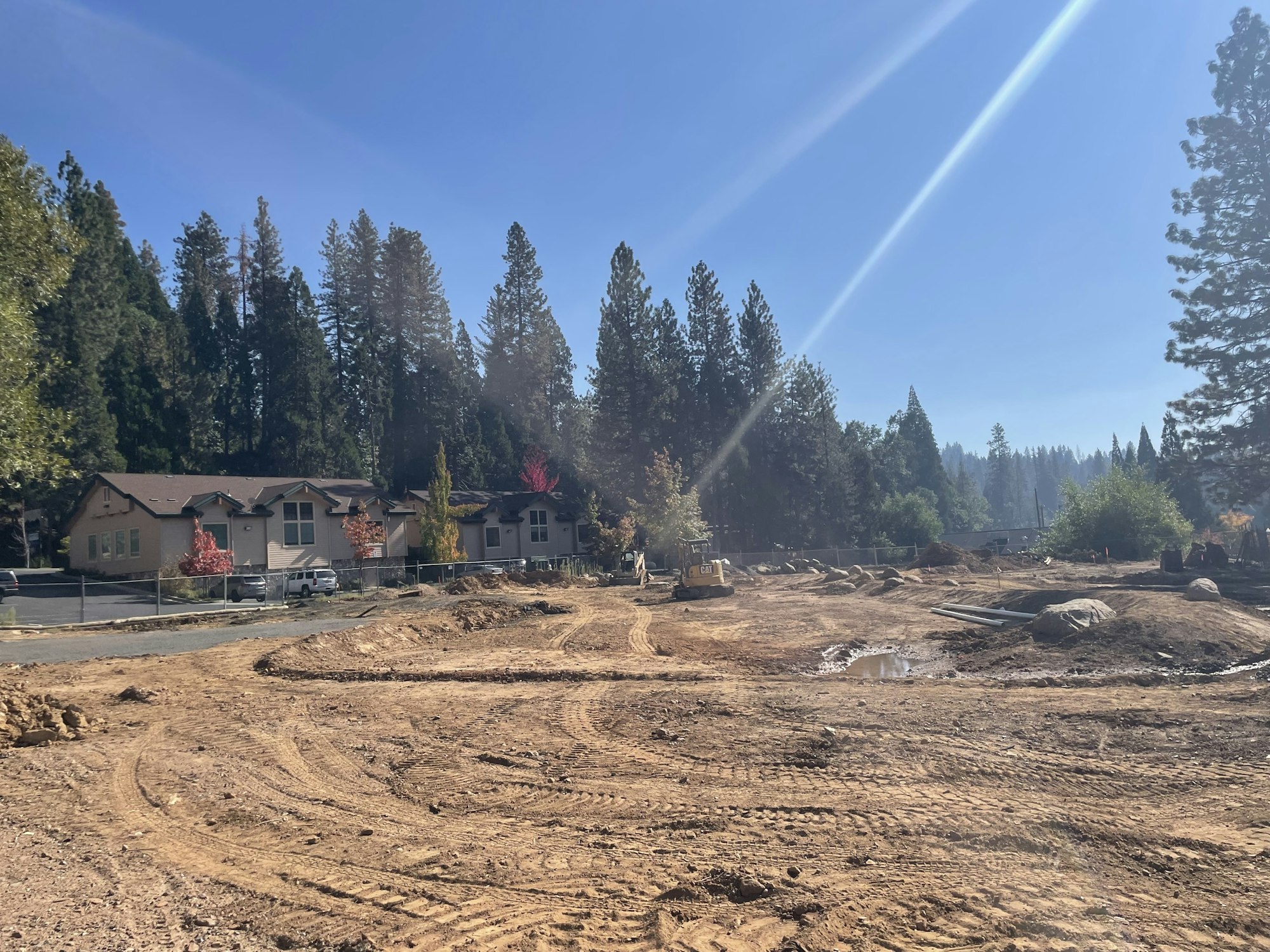 A construction site with cleared land and nearby trees, featuring a few buildings and equipment under a clear blue sky.