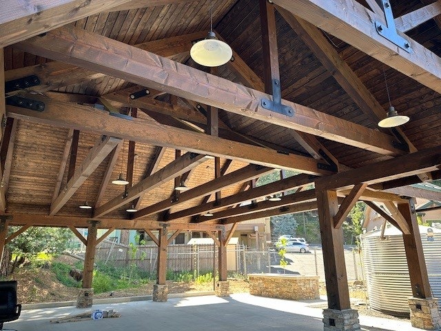 Wooden pavilion structure with exposed beams, pendant lighting, and a view to an outdoor area with a cyclist passing by.