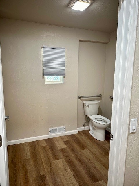 A small bathroom with a toilet, wood flooring, window with blinds, and overhead lighting.