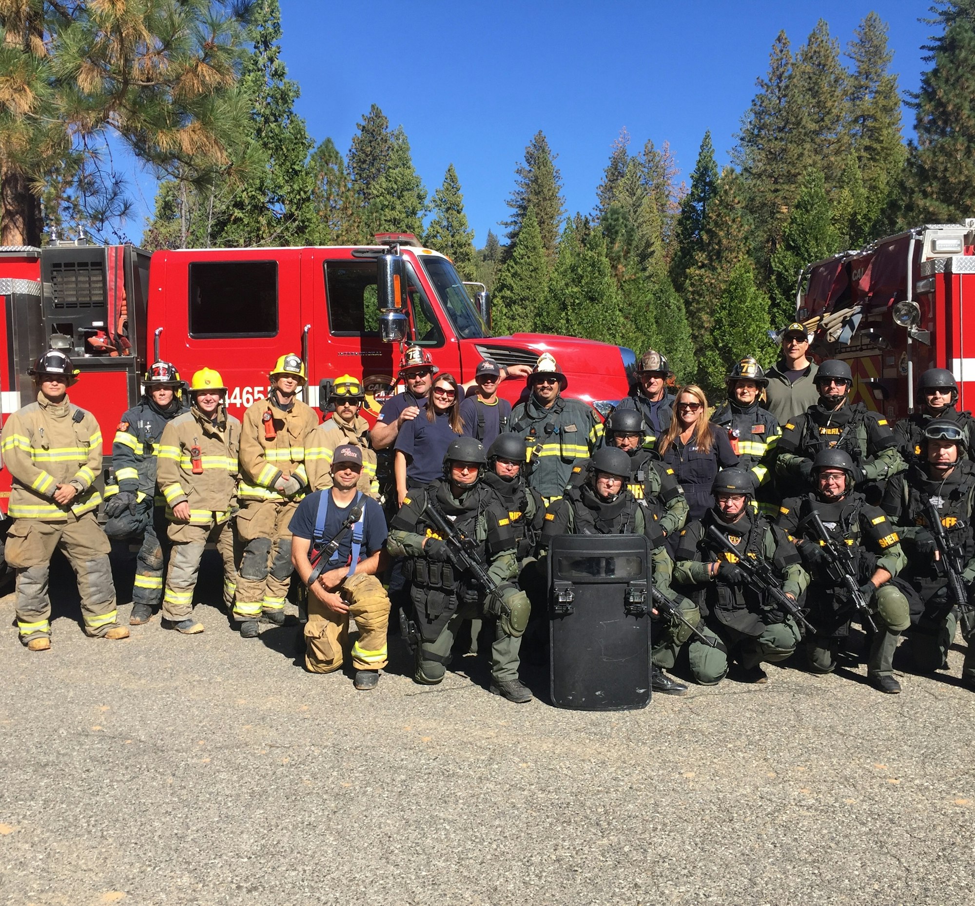A group of firefighters and law enforcement officers pose together in front of fire trucks, set in a forested area.