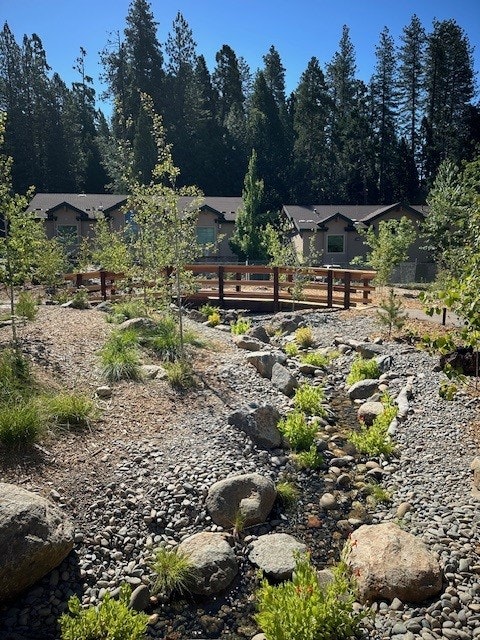 A dry creek bed with rocks and greenery, a wooden bridge, and houses among tall trees in the background.