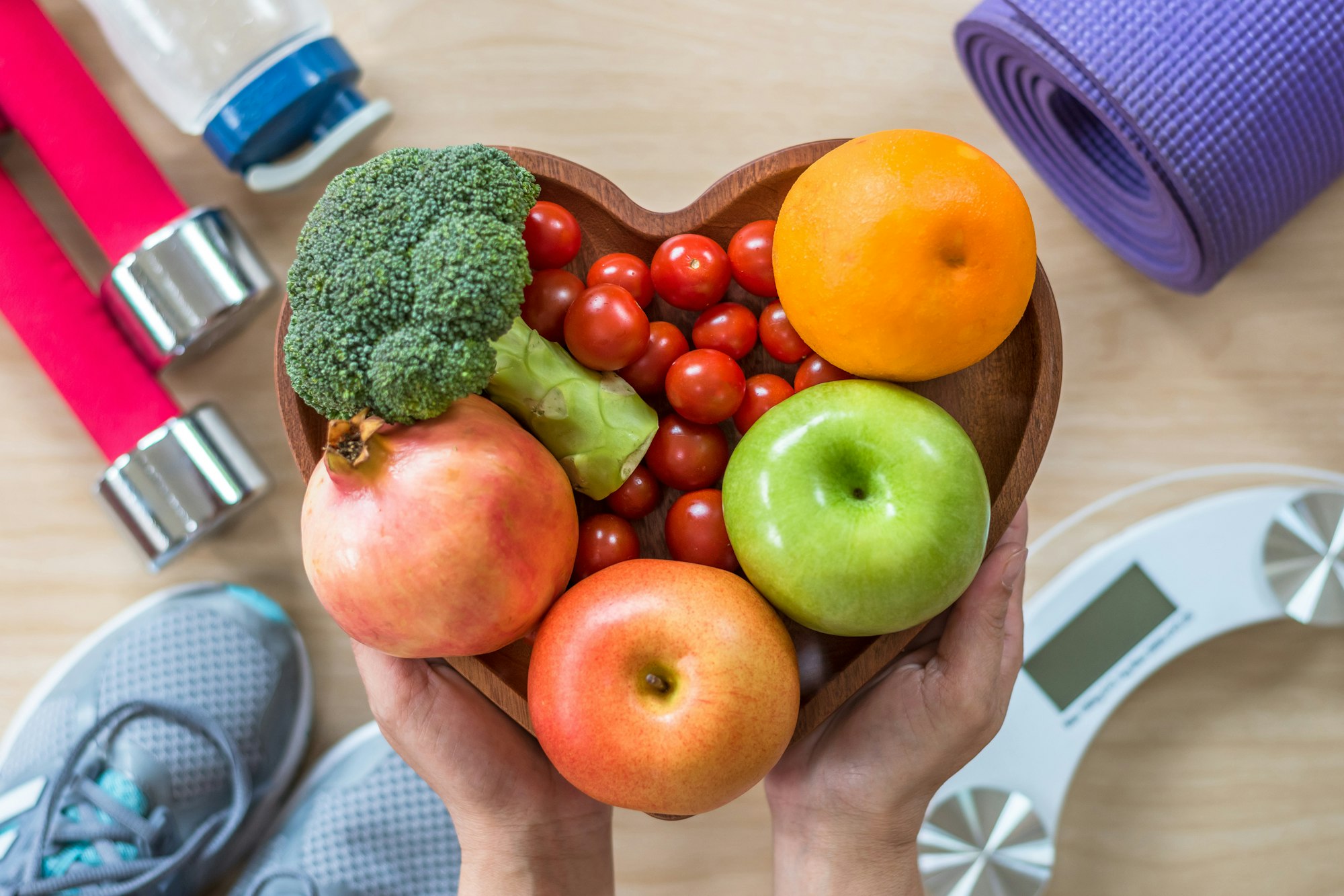 A heart-shaped bowl of fruits and vegetables held over a fitness setup with dumbbells, a yoga mat, shoes, and a scale.