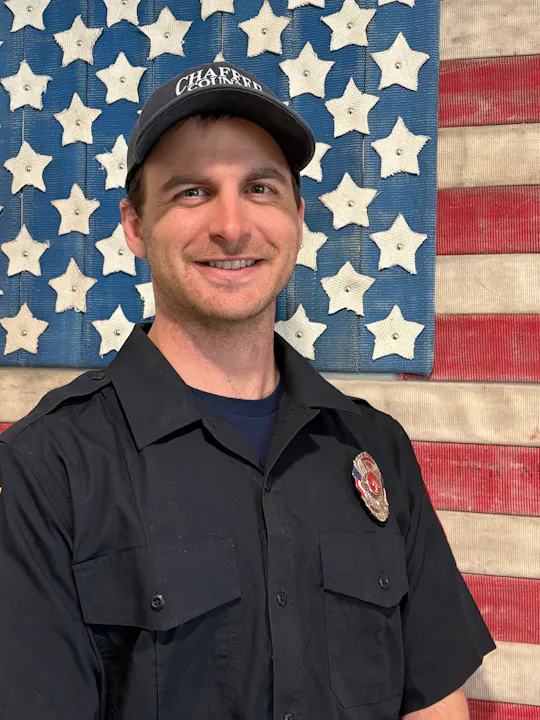 A smiling individual in a uniform stands in front of a star-patterned backdrop, resembling an American flag.
