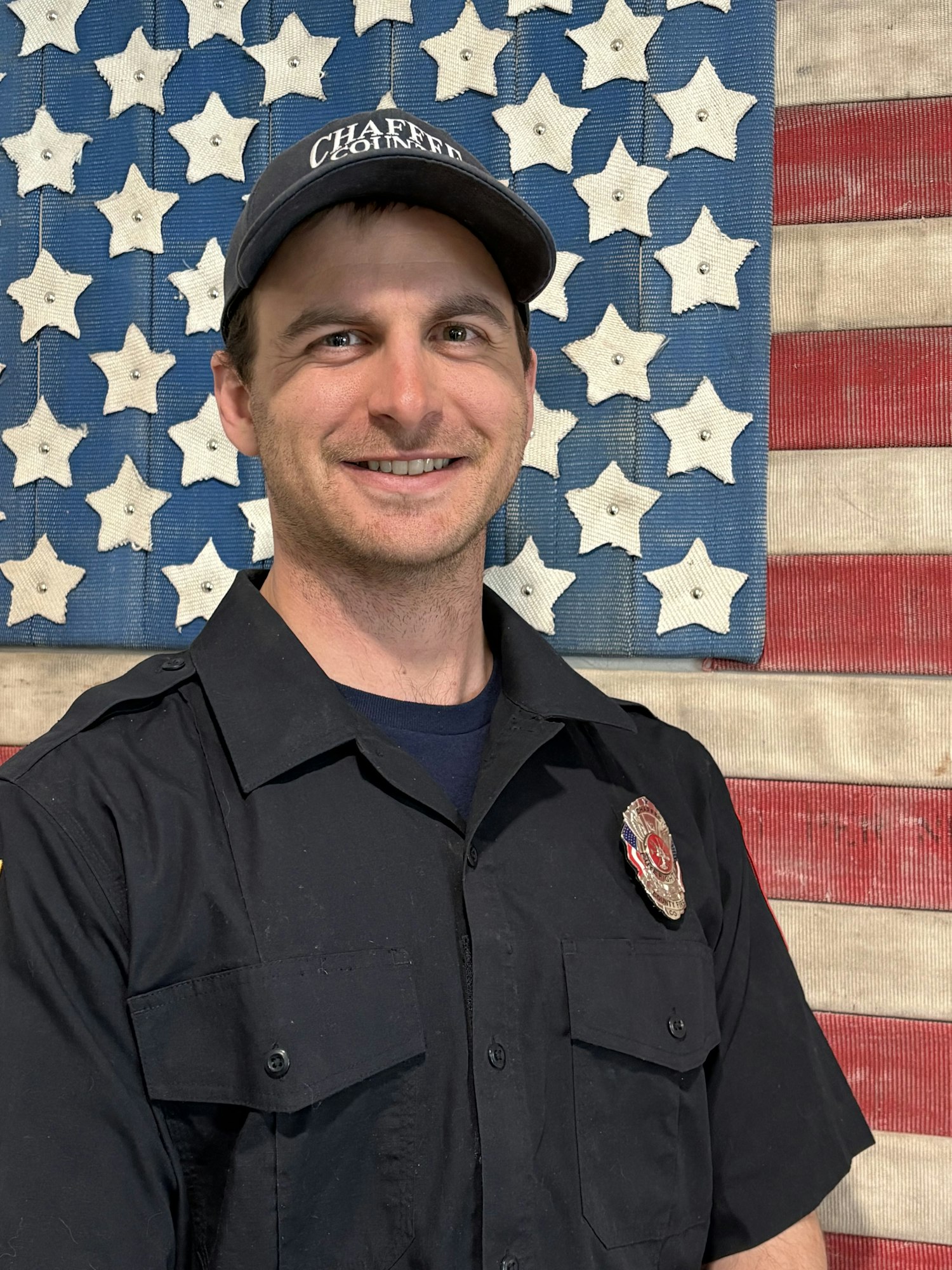 A smiling individual in a uniform stands in front of a star-patterned backdrop, resembling an American flag.