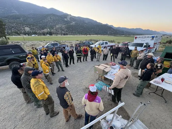 A group of emergency responders in uniforms gathers for a briefing outdoors, surrounded by vehicles and mountains.