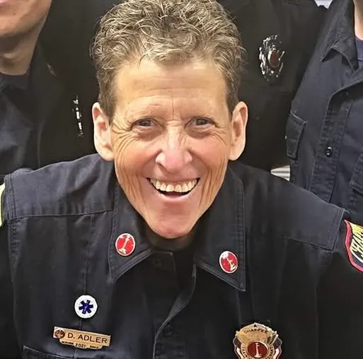 The image shows a smiling firefighter in uniform, with several badges and insignia, posing with colleagues.