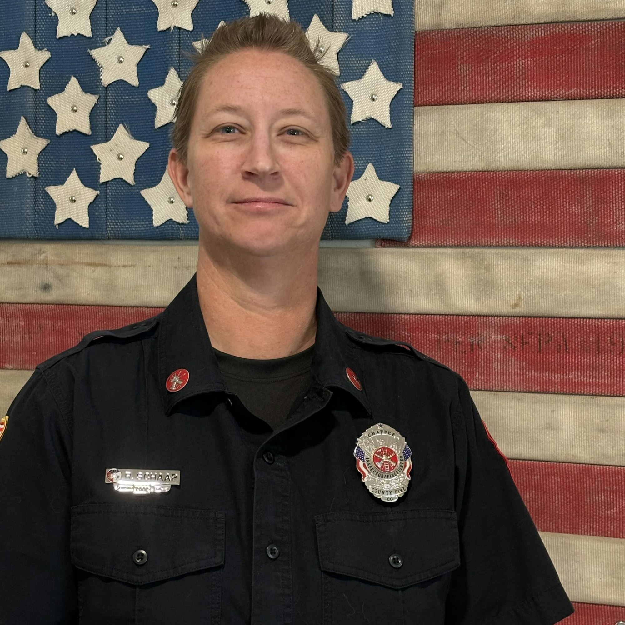 A person in a firefighter uniform stands in front of a textured American flag backdrop.