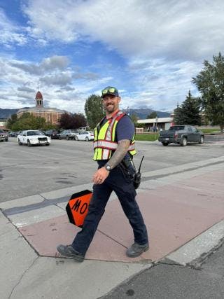 A smiling traffic officer in safety gear is walking near vehicles, holding a "MOT" (movement) sign in an urban area.