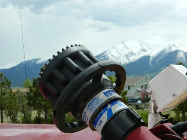 The image shows a close-up of a fire hose nozzle, with snow-capped mountains and a building in the background.