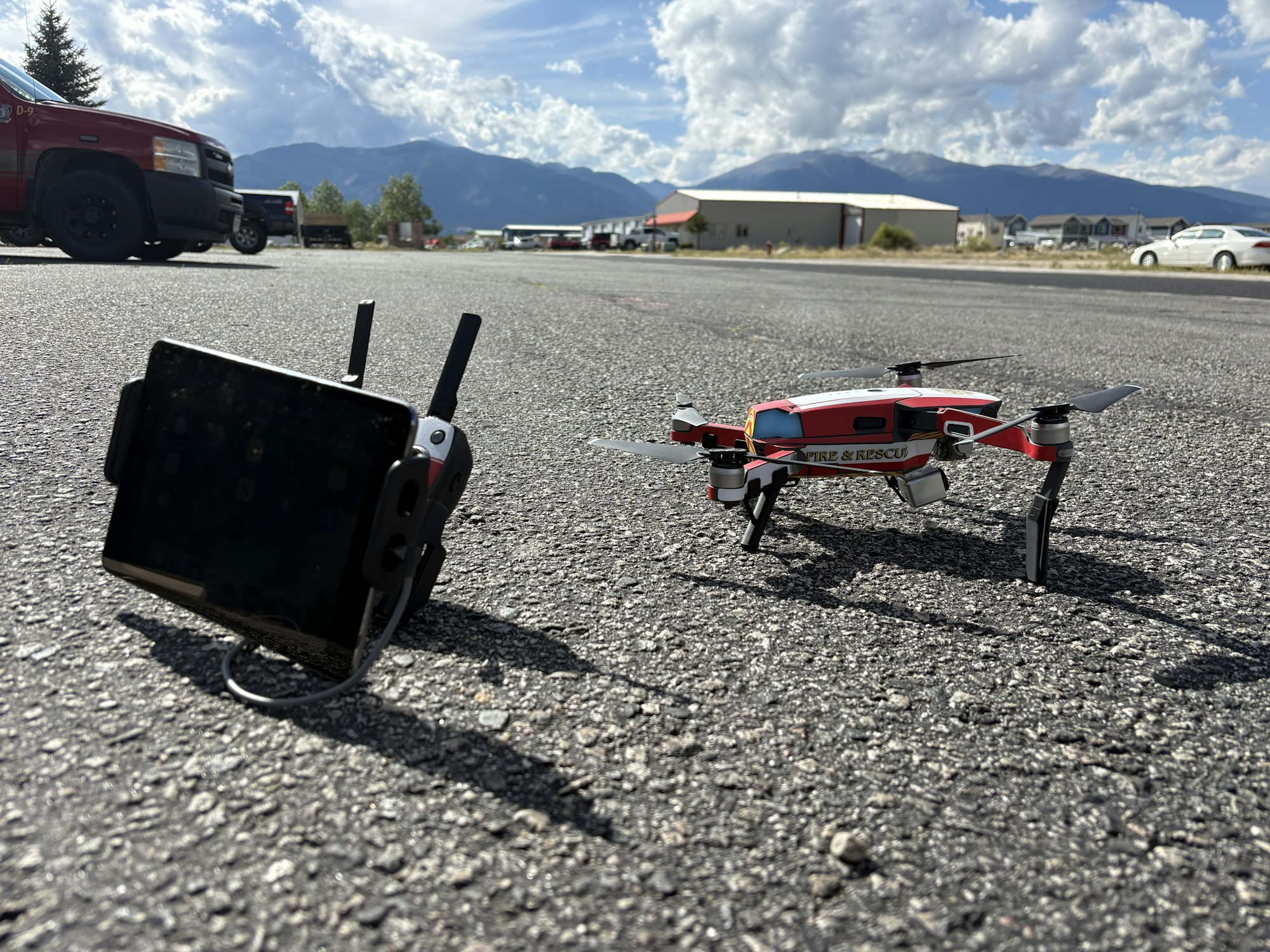 The image shows a drone labeled "Fire & Rescue" next to a remote control, set in an outdoor area with mountains in the background.