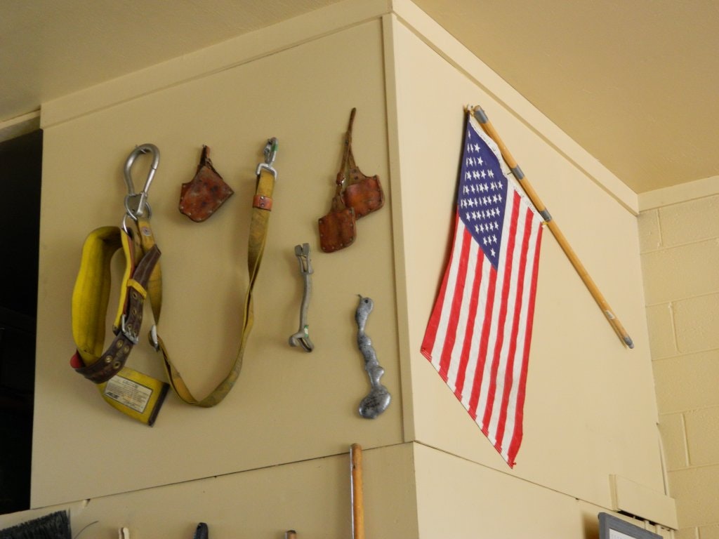 The image shows a yellow safety harness, climbing gear, metal tools, and an American flag mounted on a wall.
