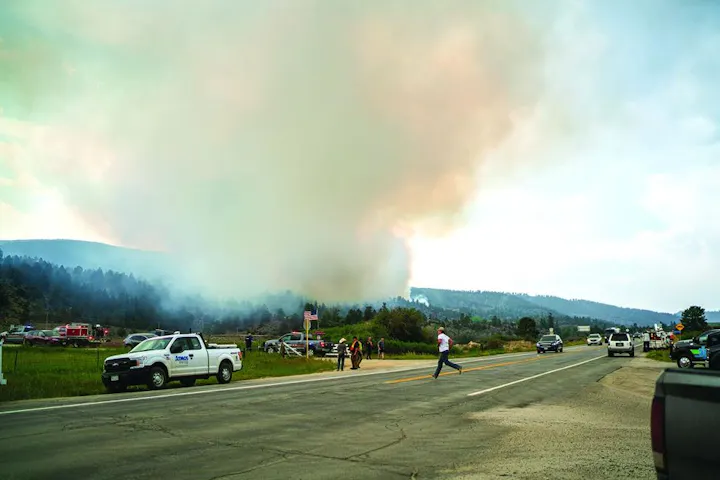 A highway scene showing smoke rising from a fire in the distance, emergency vehicles present, and people observing.