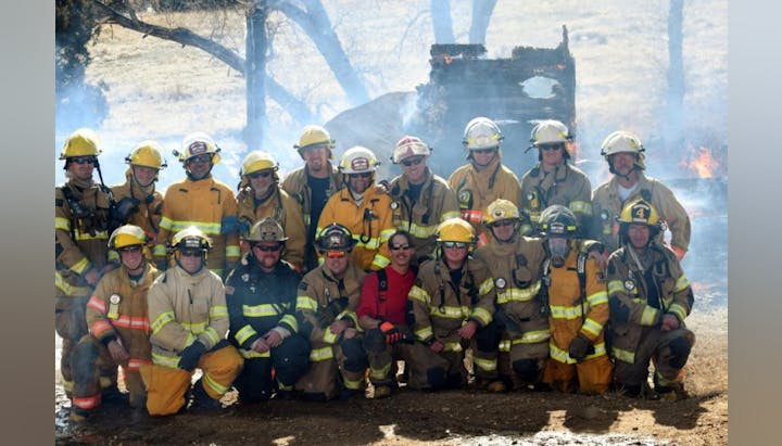 A group of firefighters poses for a photo with smoke and fire in the background, showcasing their teamwork and readiness.