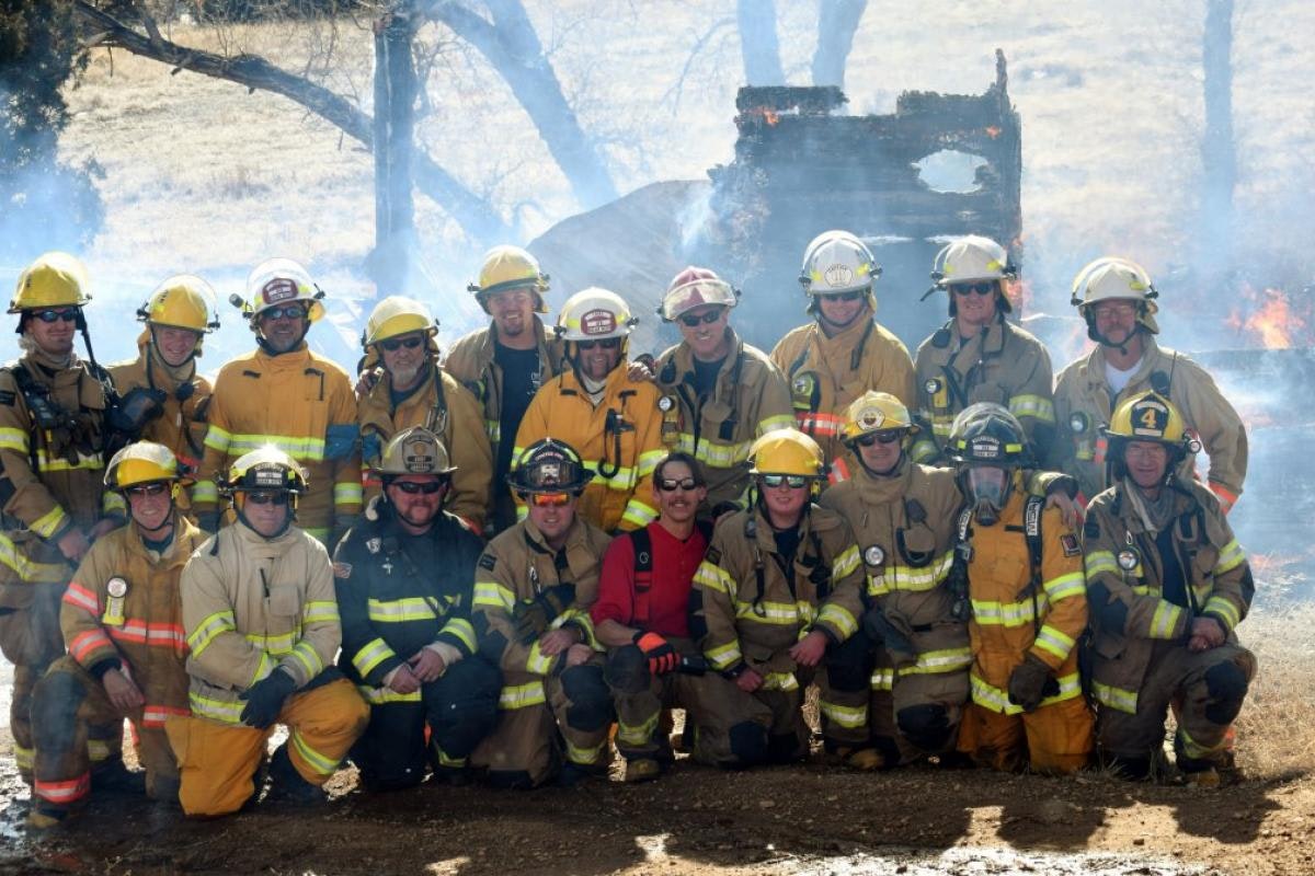 A group of firefighters poses for a photo with smoke and fire in the background, showcasing their teamwork and readiness.