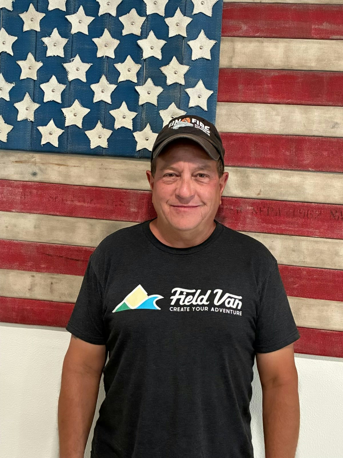 A man wearing a black "Field Van" t-shirt stands in front of a star and stripe American flag backdrop.