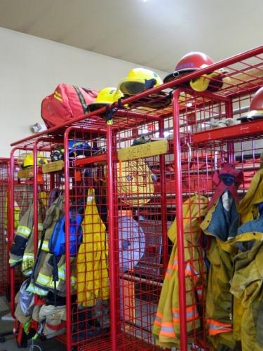 The image shows red storage racks with firefighter helmets, gear, and uniforms organized inside a fire station.
