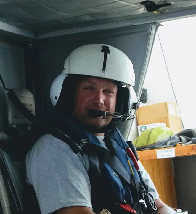 A man wearing a helmet and headset is sitting inside a helicopter, looking directly at the camera.