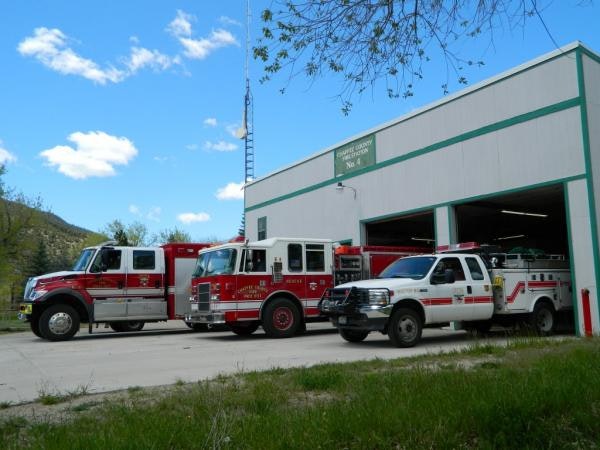The image shows a fire station with three fire vehicles parked outside, set against a blue sky with a few clouds.