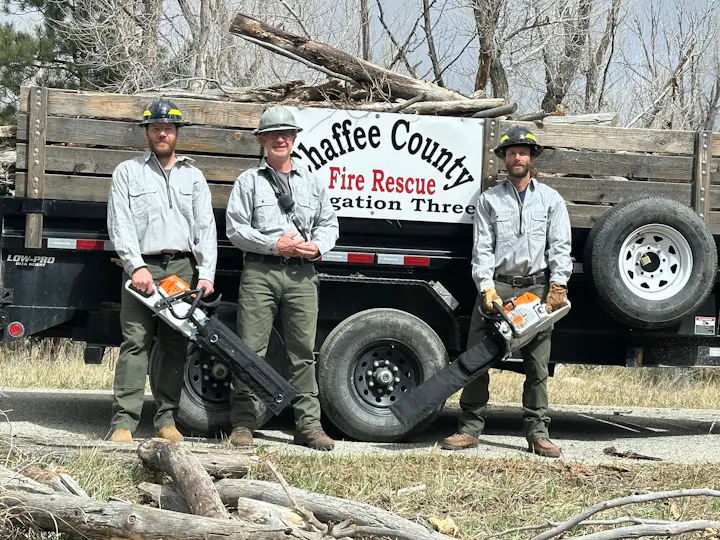 Three firefighters stand in front of a truck labeled "Chaffee County Fire Rescue," holding chainsaws in a woodland area.