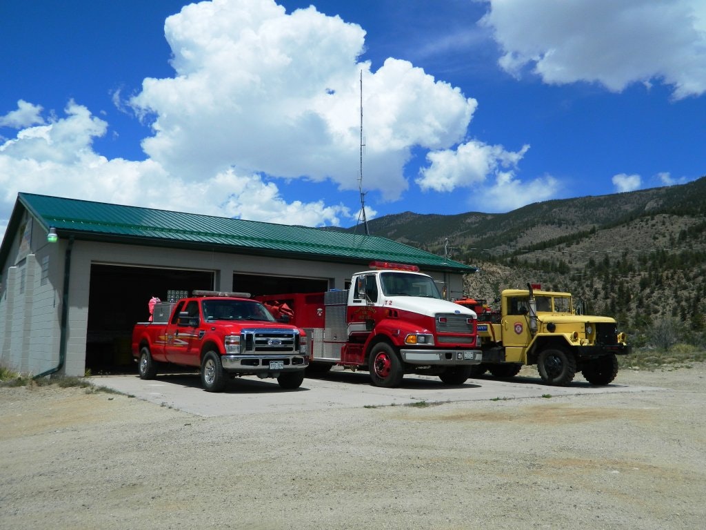 A fire station with a red fire truck, a utility truck, and a yellow vehicle, set against a mountain backdrop under a blue sky.
