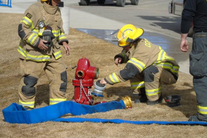 Two firefighters in gear are preparing to connect a hose to a fire hydrant on a grassy area.