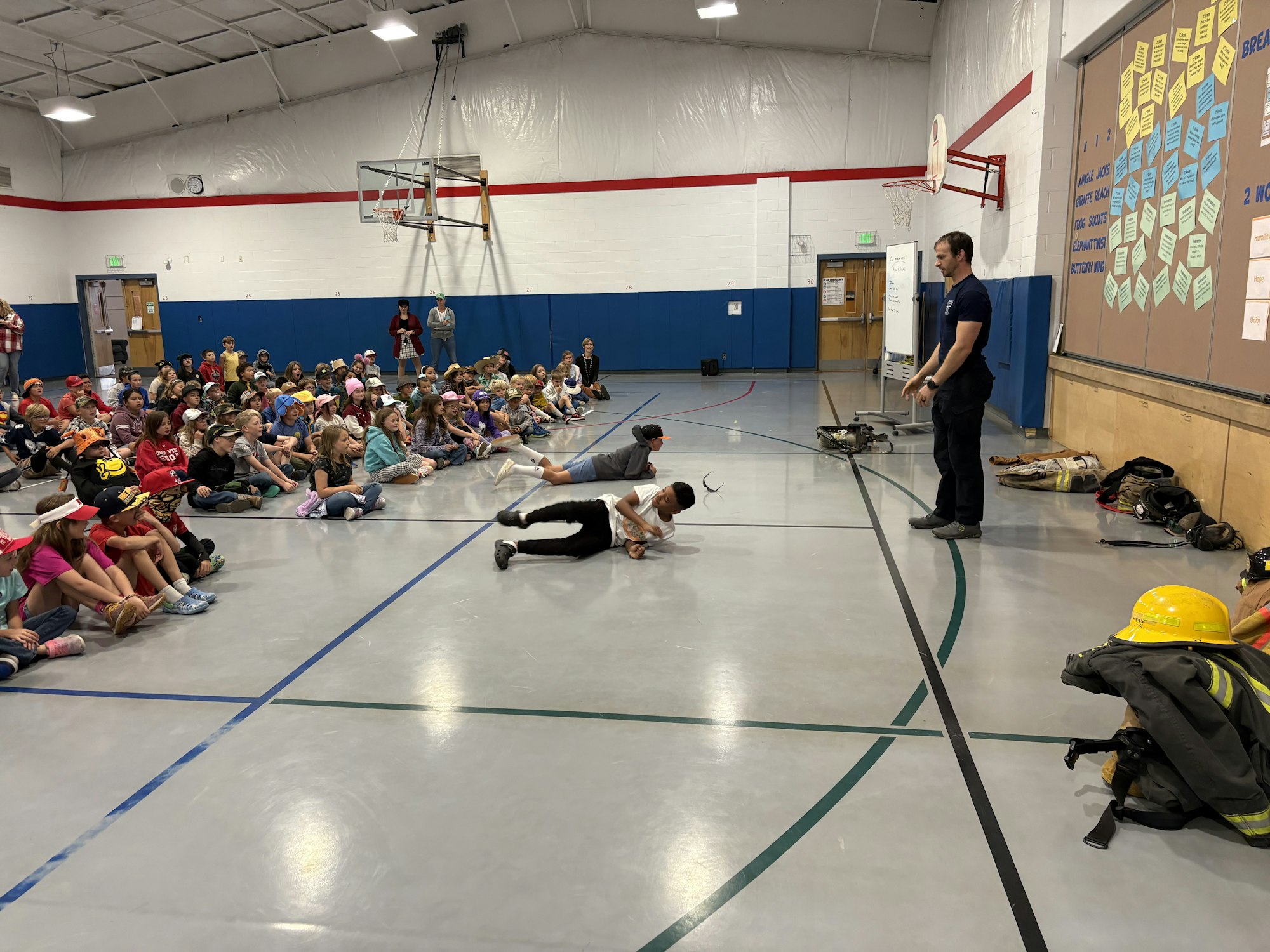 A group of children watch a presenter demonstrating activities in a gym, with some kids participating on the floor.