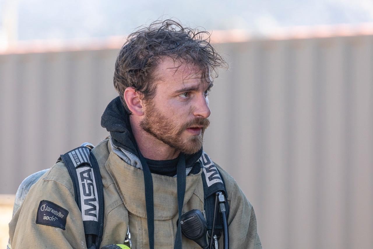 A close-up of a firefighter in gear, looking focused and serious, with a rugged appearance and textured background.