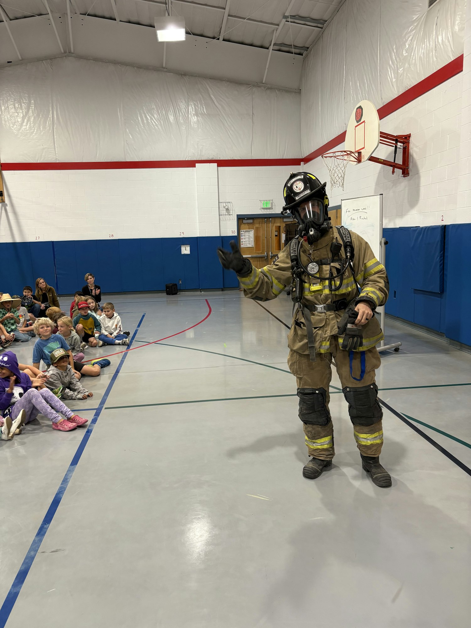 A firefighter in full gear is demonstrating safety to a group of children in a gymnasium setting.