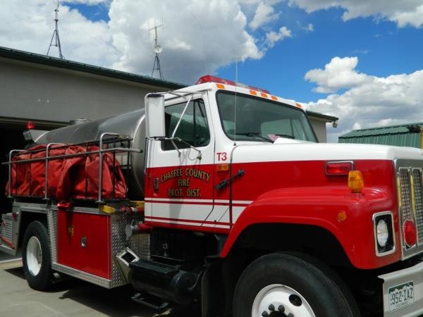 A red fire truck from Chaffee County Fire District parked outside, featuring a water tank and a blue sky with clouds.