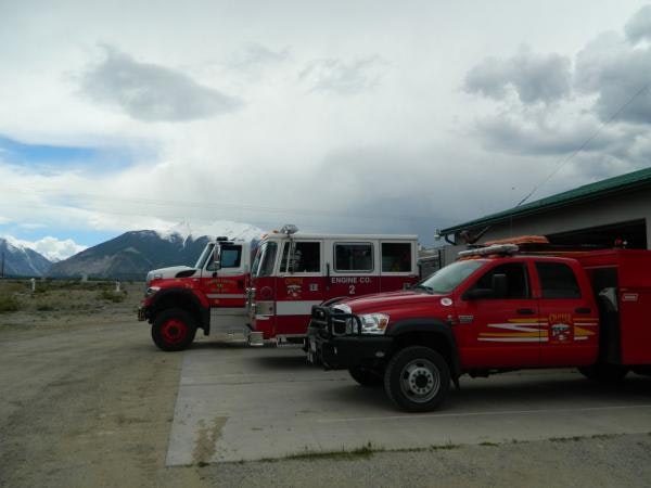 Three red fire trucks parked outside a station, with mountains and a cloudy sky in the background.